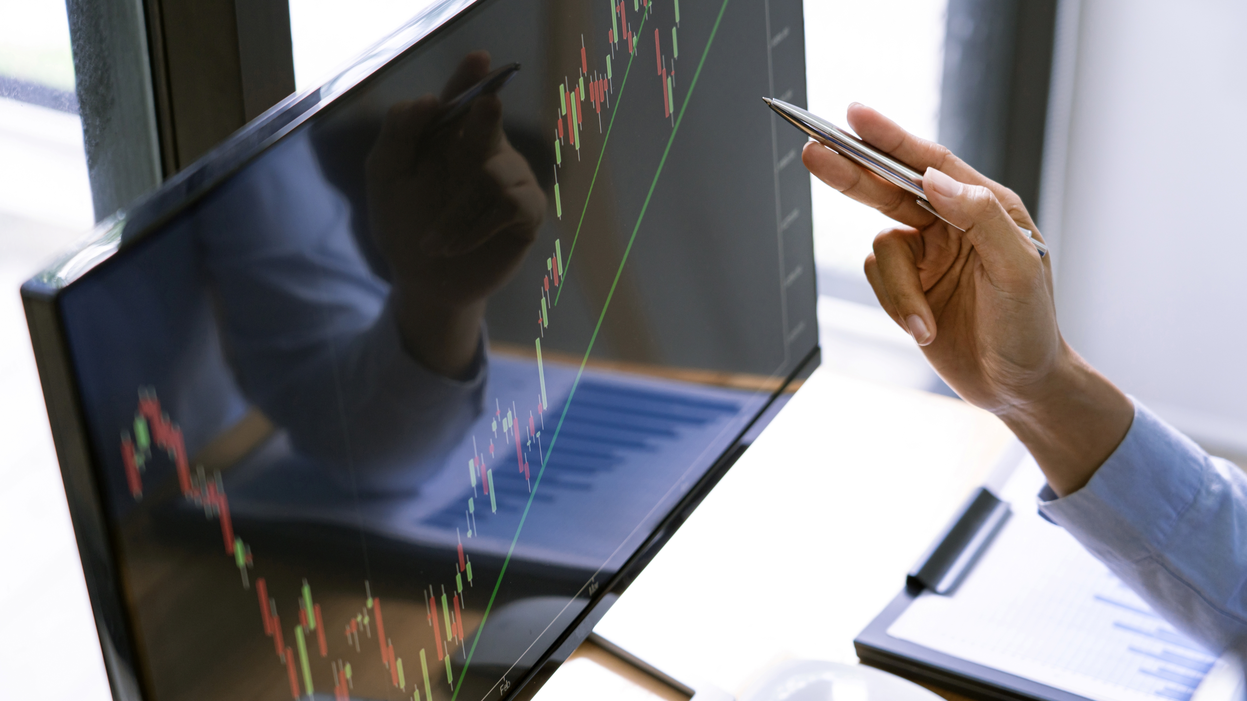 Close-up of a person holding a pen and analyzing a financial stock chart with candlestick patterns and trend lines on a computer monitor.
