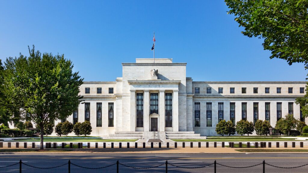 Front view of the Federal Reserve building in Washington, D.C., a neoclassical stone structure with columns, surrounded by trees under a clear blue sky.