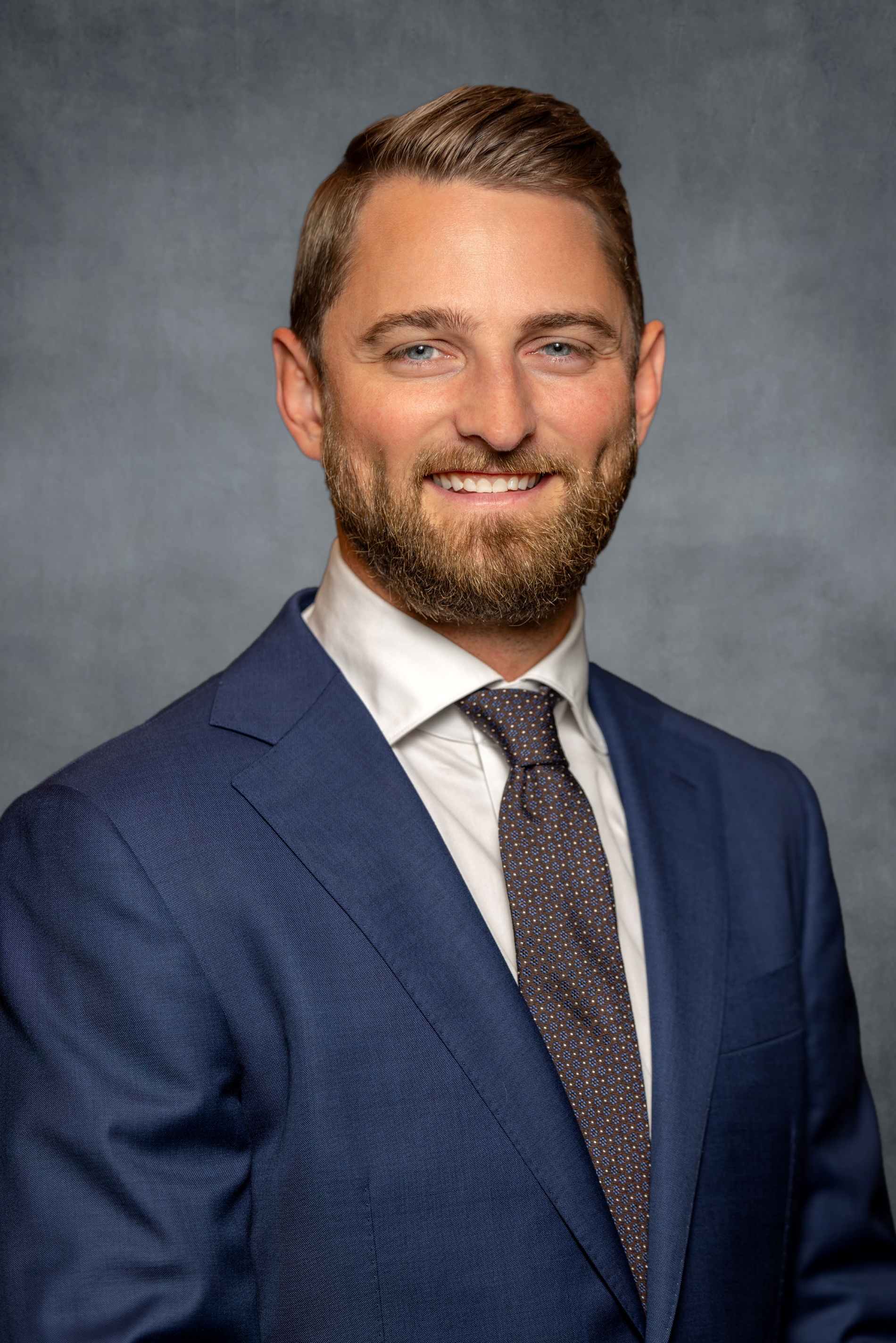 Rowen Goodman, CFP®, smiling in a professional headshot, wearing a blue suit, white dress shirt, and patterned tie against a gray studio background.